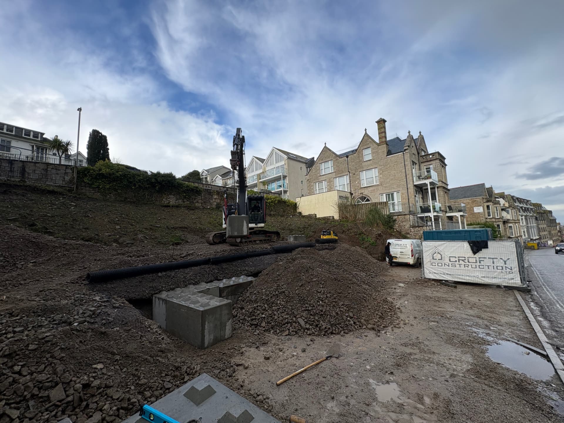 Excavator constructing a geogrid reinforced piling platform on a sloping coastal site in St Ives, Cornwall, with Crofty Construction site banner
