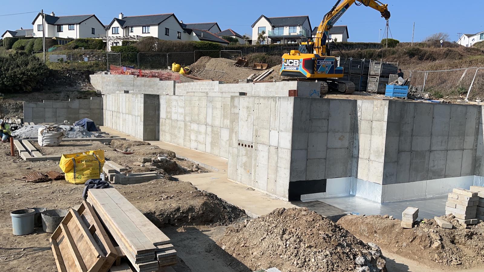 Reinforced concrete retaining walls with blockwork upstands and a JCB excavator lifting above the Porthleven site